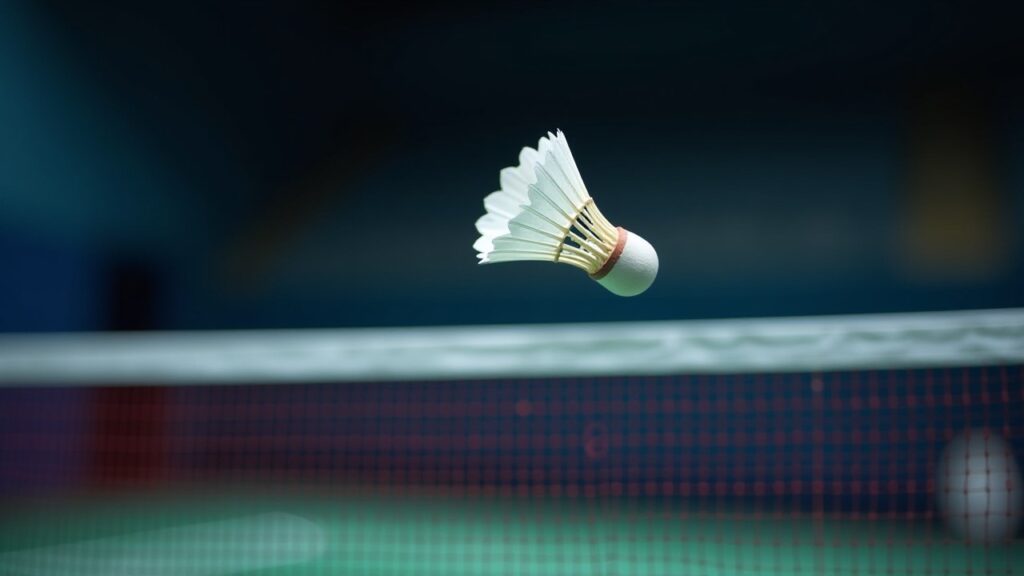 A white shuttlecock flies above the net in a dark indoor badminton hall.